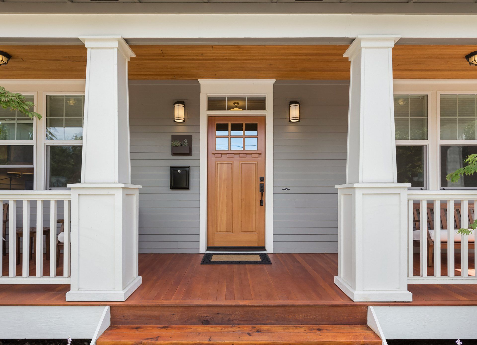 A wooden front door on a grey porch with white pillars, steps, and a natural wood ceiling.