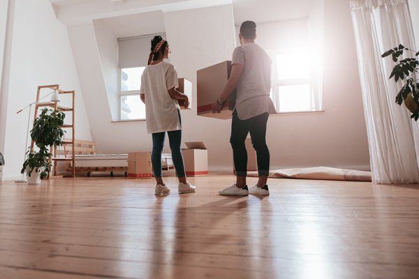 Two people stand in a bright, unfurnished room, holding cardboard boxes while moving into a new home.