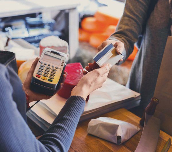 Customer Paying for Order — Charlotte, NC — AAC Insurance Group A customer hands a credit card to a cashier holding a handheld card terminal in a shop.