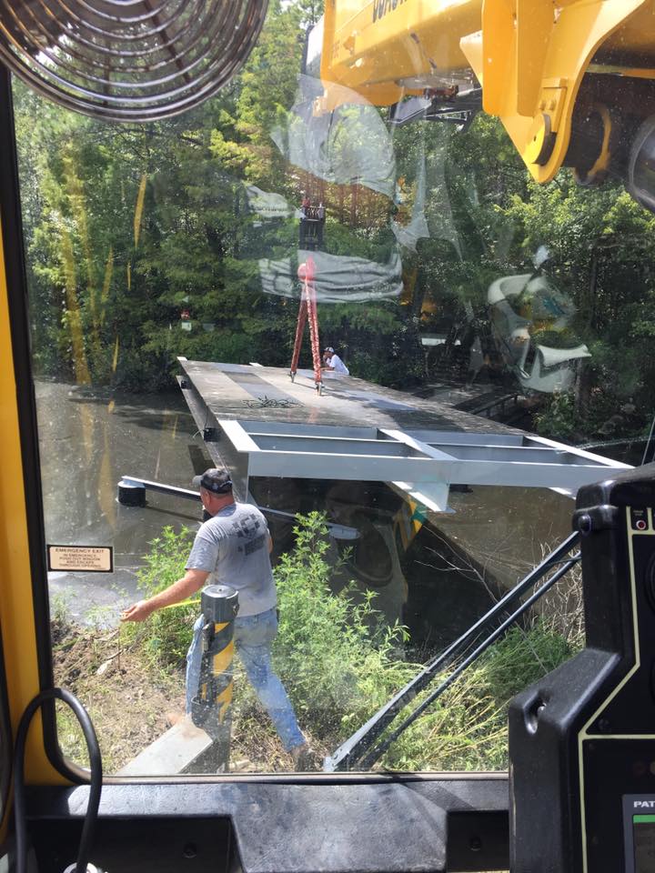 Construction worker guiding a suspended steel beam near a glass wall in a wooded outdoor setting
