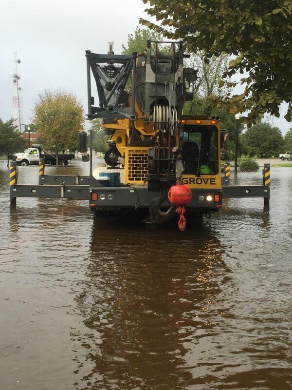 Yellow crane truck driving through floodwater on a flooded street, with a red hook hanging front center