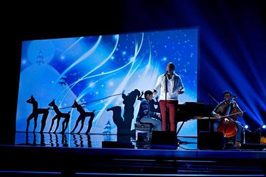 Live concert stage with blue lights, silhouetted dancers, a singer, and a seated cellist