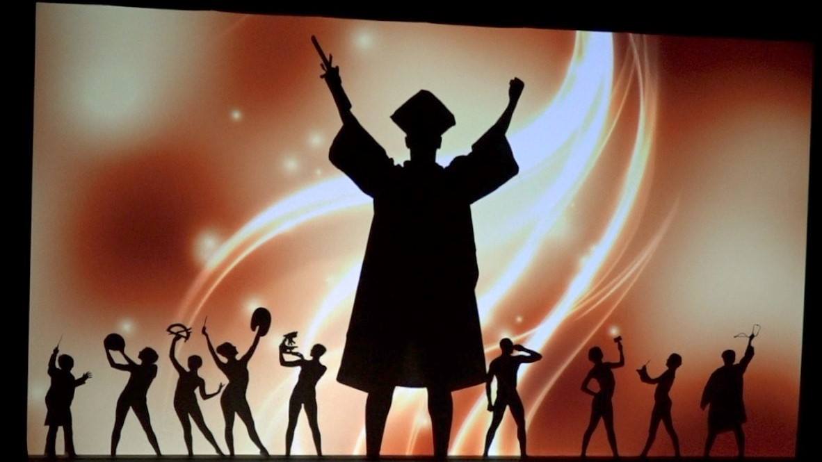 Silhouetted dancer with raised arms on stage, surrounded by cheering figures against a warm orange backdrop