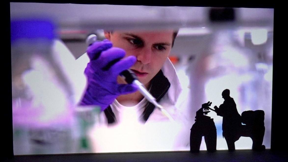 Scientist in purple gloves holding a pipette in a lab, with silhouetted workers in foreground.