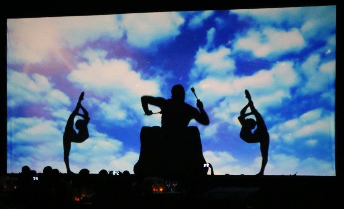 Silhouetted dancers pose against a bright blue sky with clouds on a stage