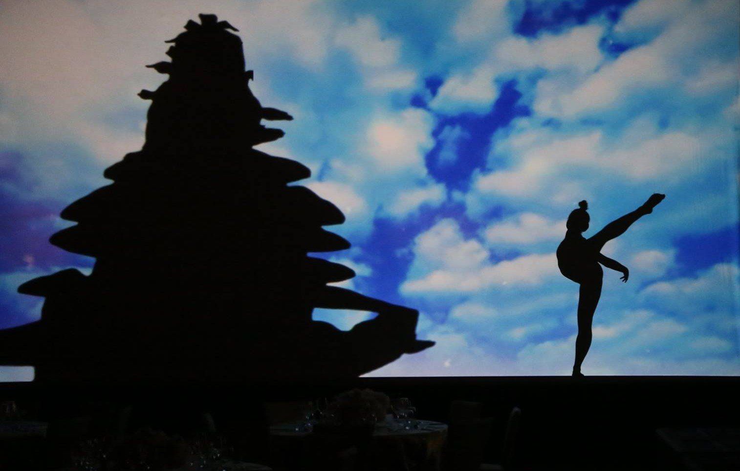Silhouetted dancer posing beside a stacked rock sculpture against a blue, clouded sky