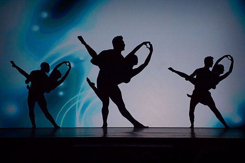 Three silhouetted dancers pose on a blue-lit stage with swirling light trails