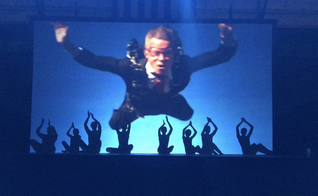 Performer on stage with arms raised, silhouetted crowd below, blue lighting and a projected figure behind.