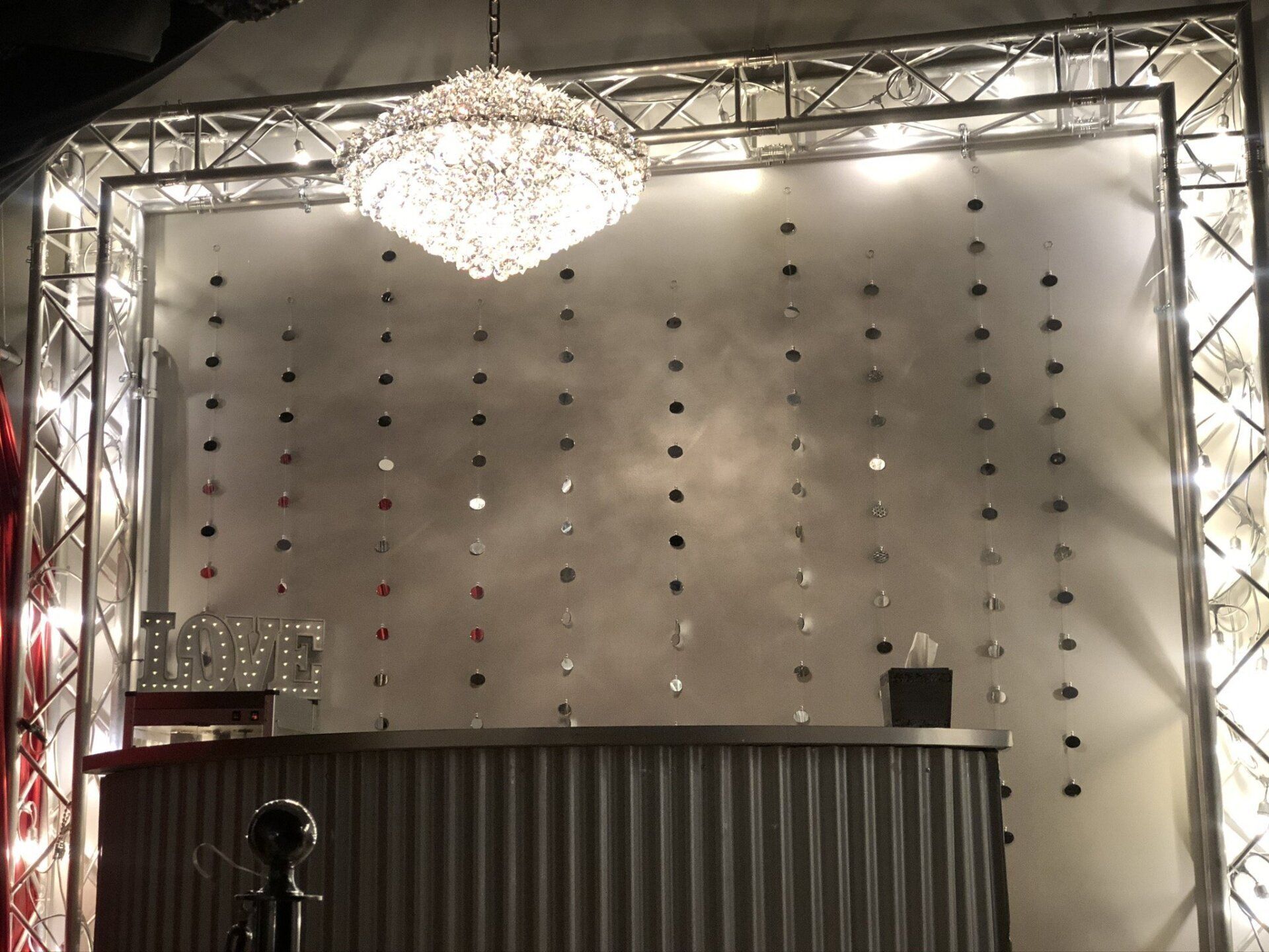 Empty event stage with a chandelier, silver backdrop, and corrugated front desk panel.