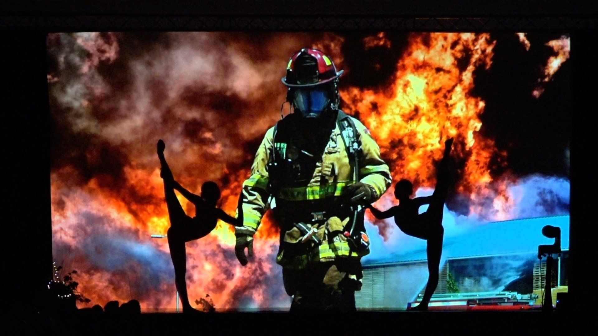 Firefighter in yellow gear standing before a blaze, with flames and smoke filling the background.
