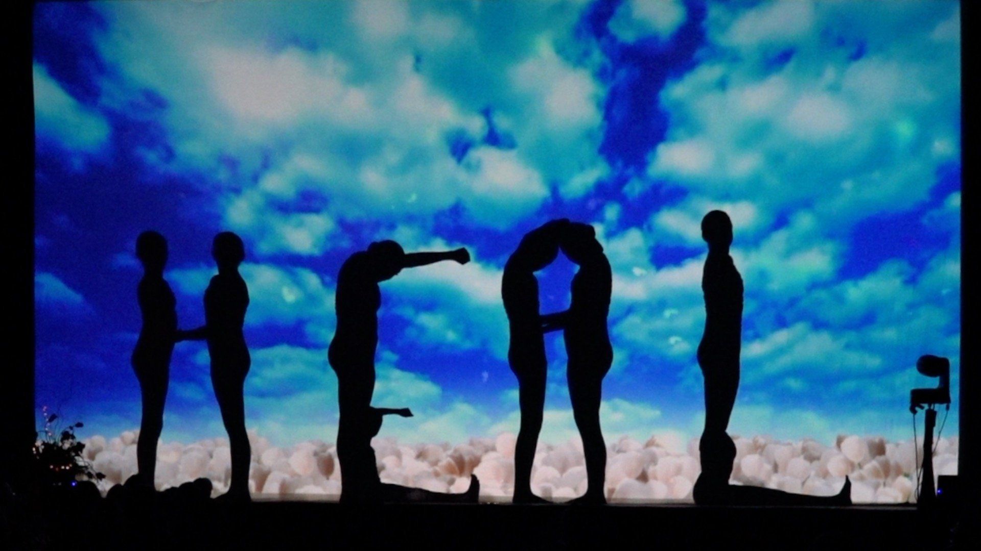 Silhouetted people spell “HELLO” against a bright blue cloudy sky.
