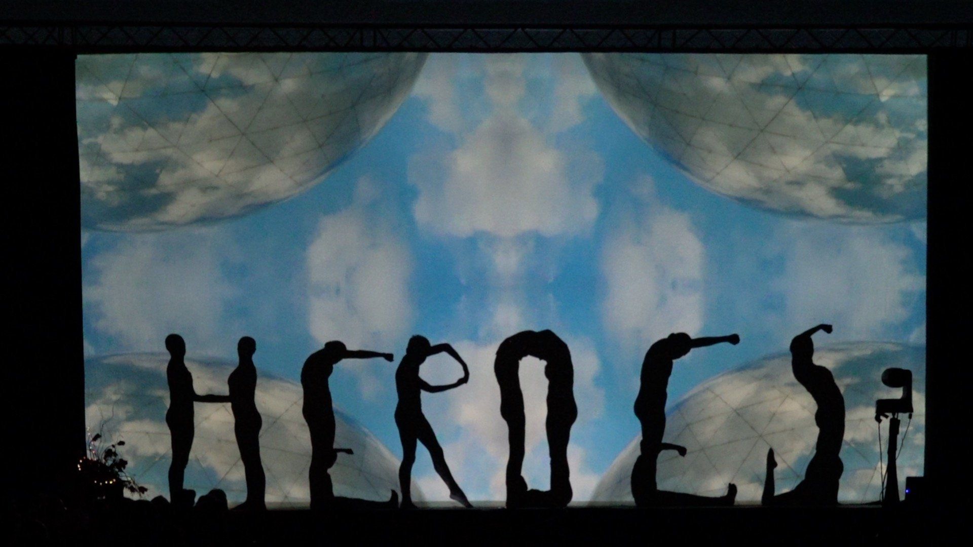 Silhouetted people forming letters against a cloudy blue sky and horizon.