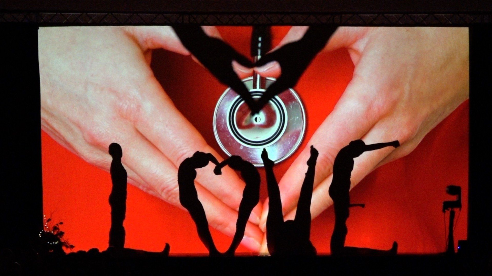 Silhouetted people spelling “LOVE” against a red background with a heart shape above them
