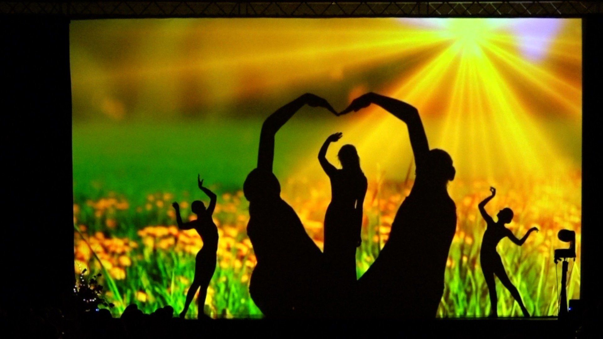 Silhouetted people celebrating in a sunlit field of yellow flowers, with a heart shape formed by raised arms