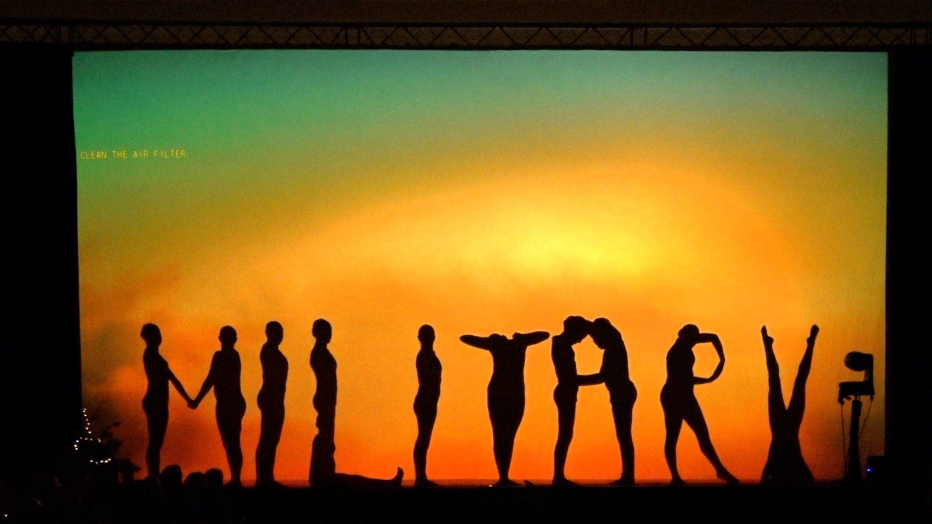 Silhouetted people forming “MILITARY” against a glowing orange sunset backdrop.