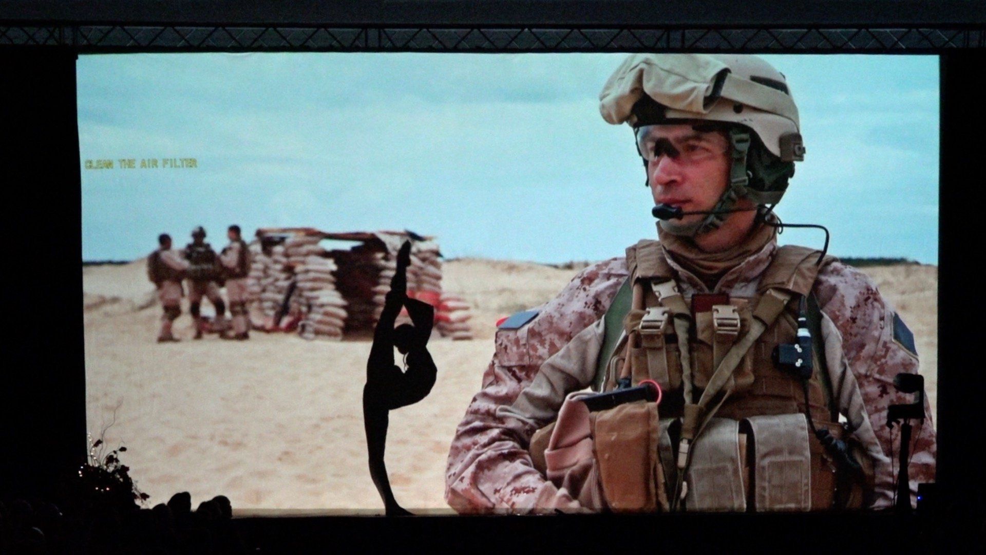 Soldier in desert camouflage stands guard near a sandbag wall in a barren landscape.