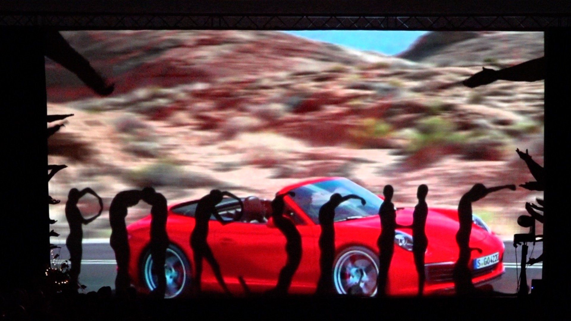 Red sports car on stage with silhouetted people in front of a large scenic backdrop