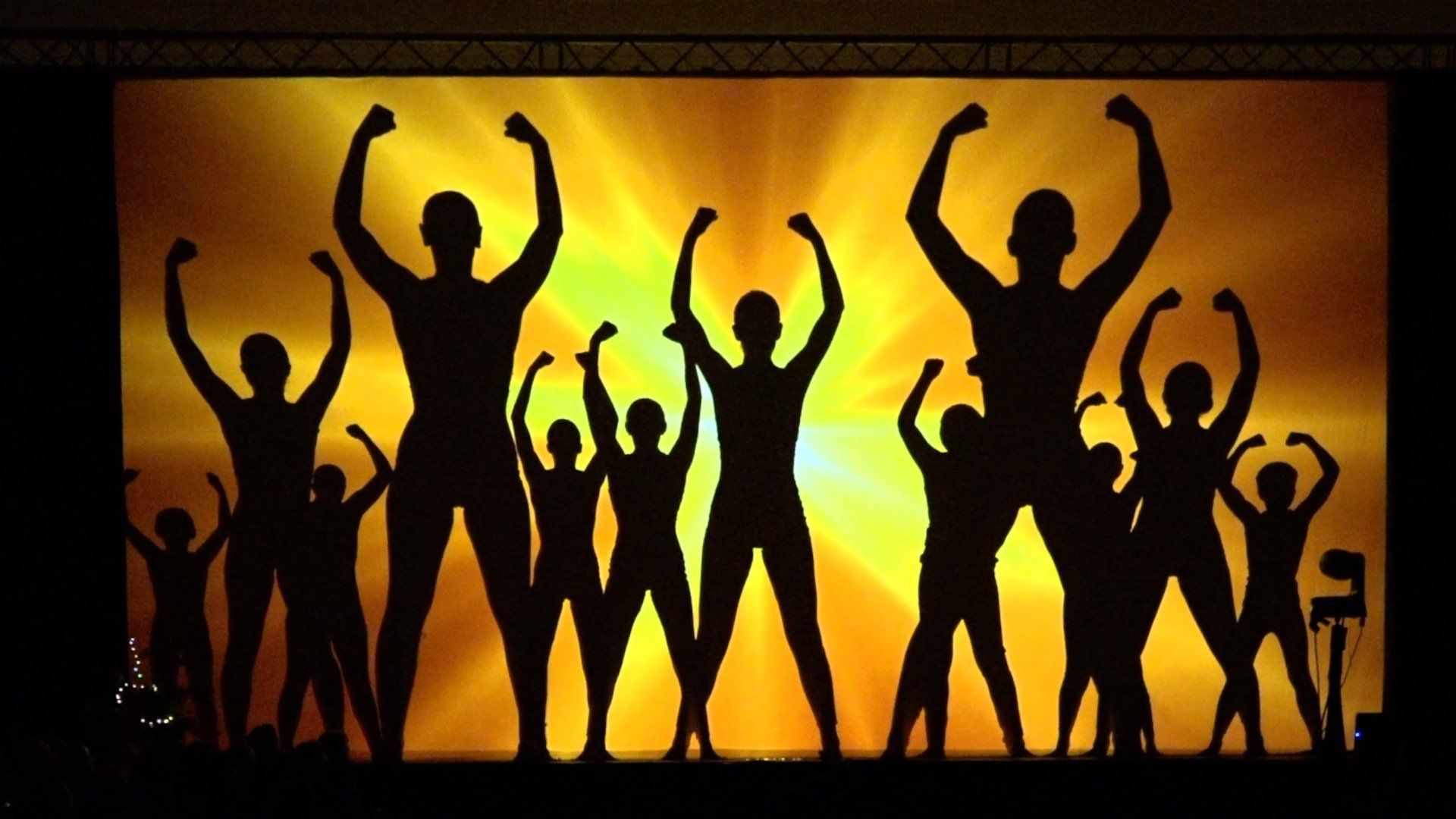 Silhouetted people dancing with raised arms against a glowing orange and yellow stage backdrop