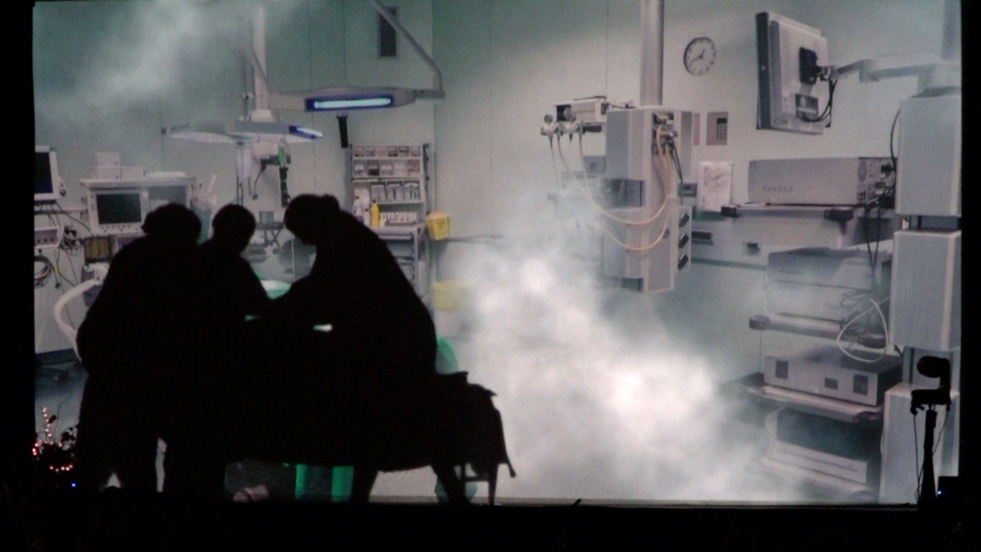 Silhouetted medical staff in a misty hospital room with monitors and equipment