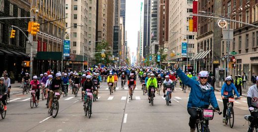 Cyclists ride down a city street during a bike event, with one rider in blue waving in the foreground.