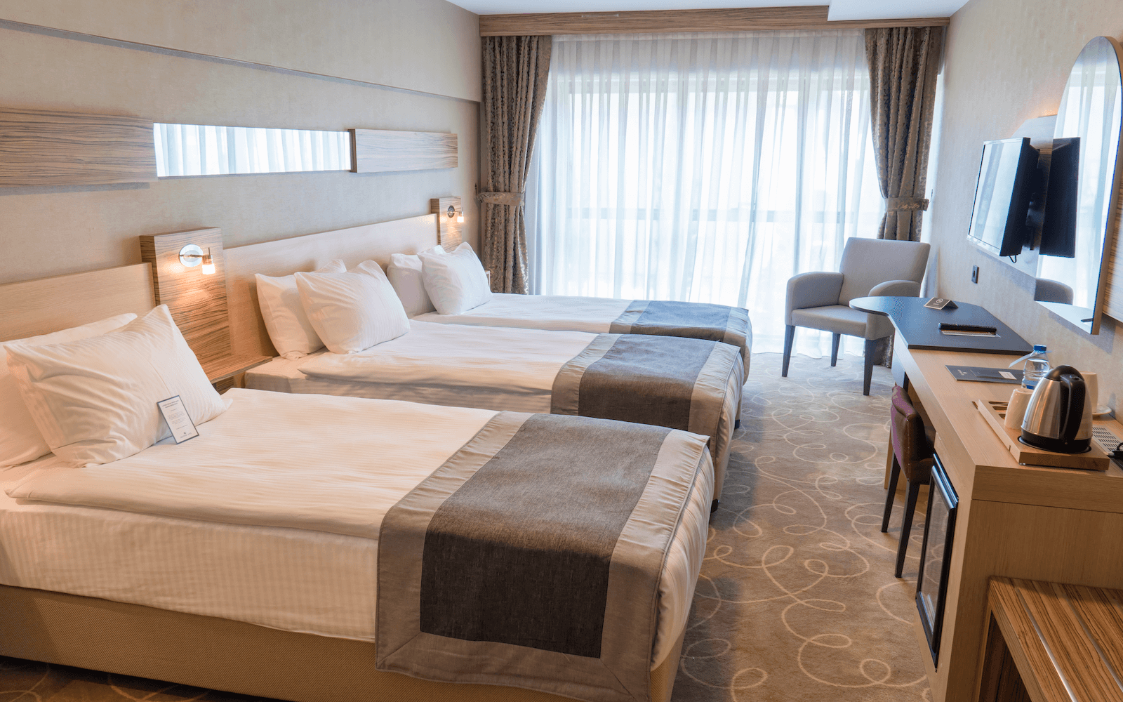 Üç yataklı, masalı, sandalyeli ve televizyonlu bir otel odası.