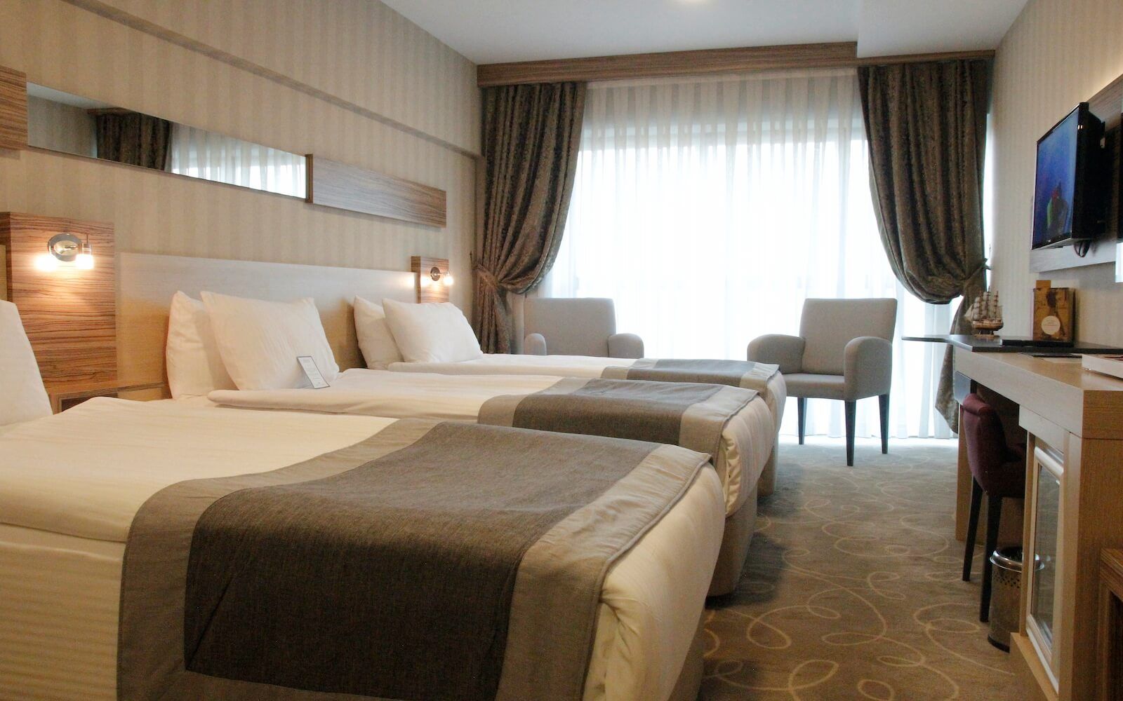 Üç yataklı, masalı, sandalyeli ve televizyonlu bir otel odası.