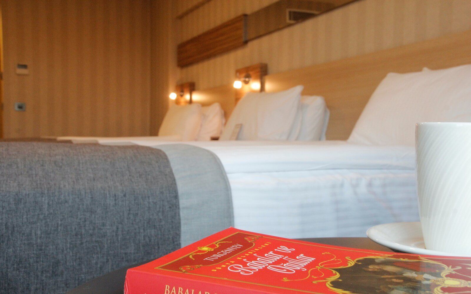 Bir otel odasındaki masanın üzerinde bir kitap ve bir fincan kahve var.