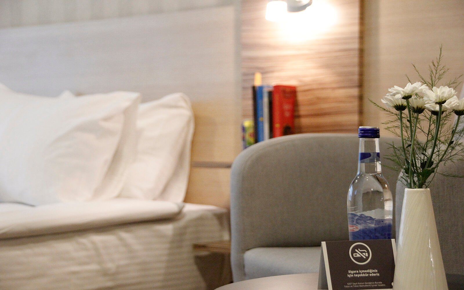 Bir otel odasındaki masanın üzerinde bir şişe su ve bir vazo dolusu çiçek var.
