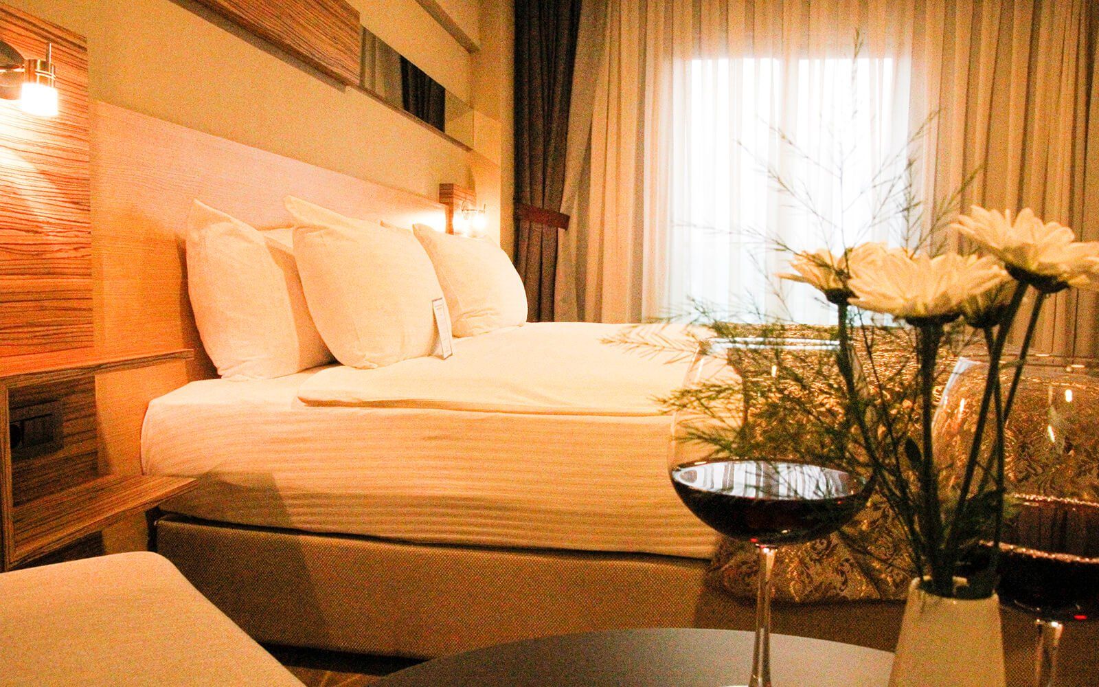 Bir yatak, bir vazo dolusu çiçek ve bir kadeh şarap bulunan bir otel odası.