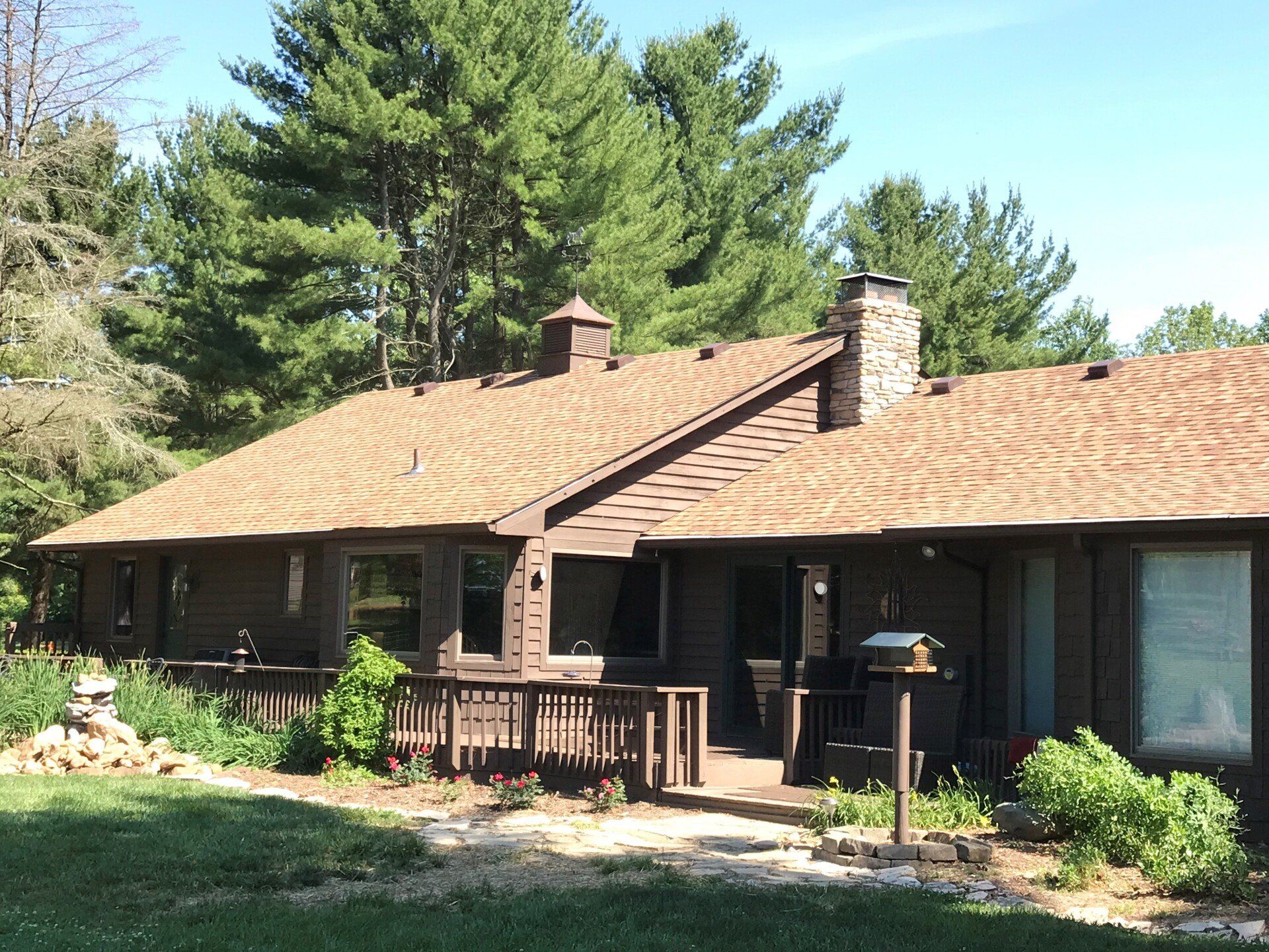 Brown house with brown shingle roof, chimney, deck, and surrounding greenery.