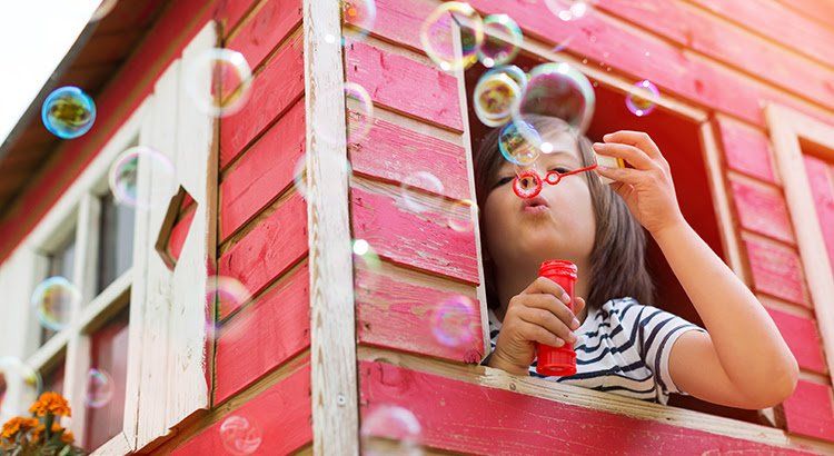 A little girl is blowing soap bubbles out of a window in a playhouse.
