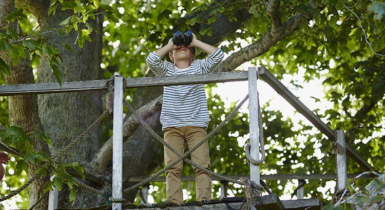 A boy is standing on a tree house looking through binoculars.