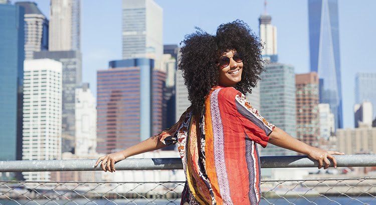 A woman with curly hair is standing on a bridge in front of a city skyline.