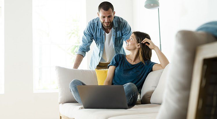 A man is standing next to a woman sitting on a couch using a laptop computer.