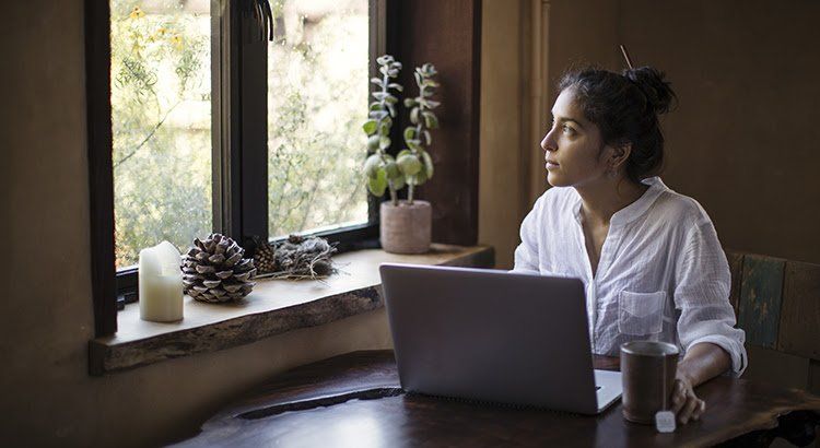 A woman is sitting at a table with a laptop and looking out the window.
