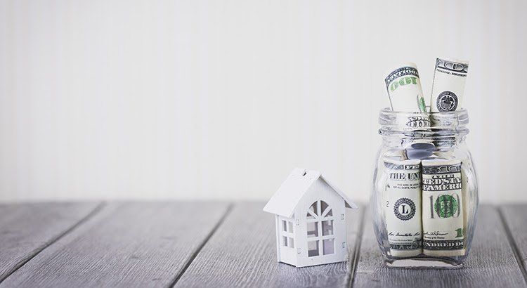 A jar filled with money and a model house on a wooden table.