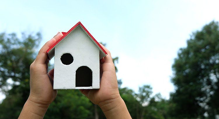 A person is holding a small white house with a red roof.