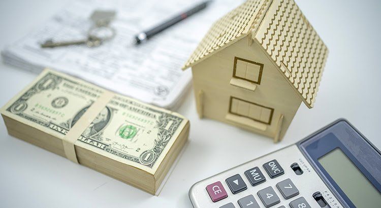 A stack of money , a model house , and a calculator are on a table.
