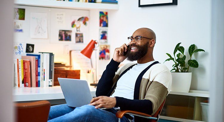 A man is sitting in a chair using a laptop and talking on a cell phone.