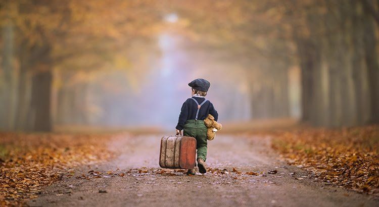 A young boy is walking down a road with a suitcase and a teddy bear.