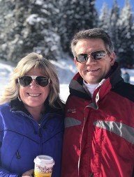 Couple smiling in front of a snowy background, wearing sunglasses and winter jackets.