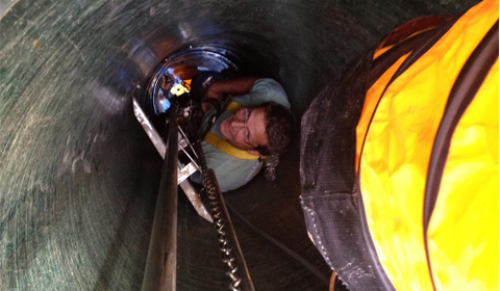 Person inside a tunnel, looking up. Wearing a harness and light. Yellow and black air duct visible.