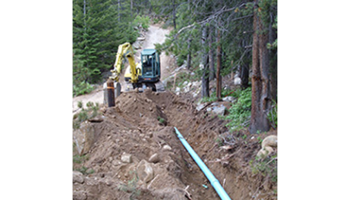 An excavator digs a trench in a forest, laying a blue pipe for construction.
