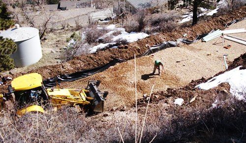 Yellow backhoe excavating earth near a man in work clothes, with a water tank and buildings in the background.
