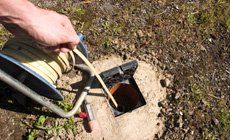 Hand with a wooden stick inspecting a drain opening near a coiled hose on the ground.