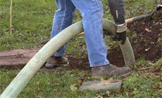 Person in blue jeans and work boots holding a large, green hose near a brown soil patch outdoors.