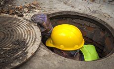 Person in a yellow hard hat and safety vest, working in a manhole, lifting the cover.