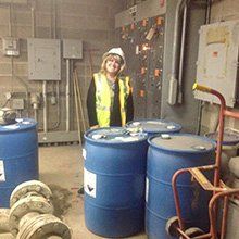 Woman in safety vest and hard hat stands in room with blue barrels, industrial setting.