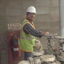 Man in hard hat and safety vest operates valve near a fire extinguisher in a utility area.
