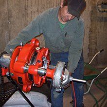 Man in a baseball cap using a large orange pipe threading machine in a basement.